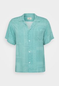 Camisa de manga curta, com gola, em tecido azul claro, apresentando um padrão subtil com manchas e texturas. Um bolso no peito.
