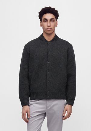 Jonge man met krullend zwart haar, gekleed in een donkergrijze vest met knopen en lichtgrijze broek, staand tegen een eenvoudige lichte achtergrond.