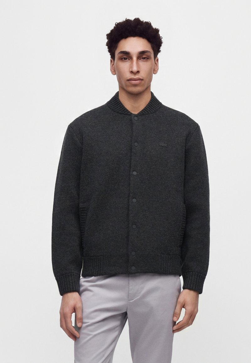 Jeune homme aux cheveux bouclés noirs portant un cardigan gris foncé boutonné et un pantalon gris clair, debout devant un fond clair uni.