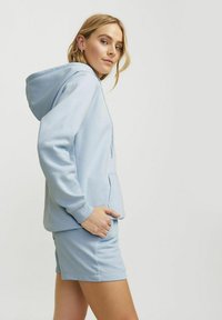 Haut à capuche bleu clair avec une poche kangourou, des manches longues et un cordon de serrage, associé à un short assorti de la même couleur et du même matériau.