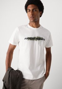 Hvit bomull t-skjorte med et svart og grønt spraymalt "ANTONY MORATO"-logo. Modellen holder en mørkegrå jakke.
