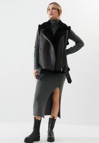 Czarna kamizelka z ekoskóry z kołnierzem z owczej wełny, zapinana na zamek, z bocznymi paskami, noszona na szarej dopasowanej sukience z rozporkiem do uda oraz czarnych botkach.