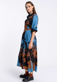 Długa sukienka w niebiesko-brązowo-czarny abstrakcyjny wzór, z guzikami z przodu i związanym w talii brązowym paskiem. Gładka tekstura tkaniny.