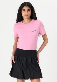 T-shirt en coton rose à manches courtes, comportant un texte en cursive noir "Daisy" sur le côté gauche. Associé à une jupe noire froncée.
