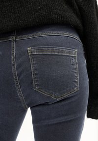 Jeans azul oscuro de corte slim, con costuras amarillas, un pequeño bolsillo en la parte trasera y una textura de denim suave. Diseño de cintura alta.