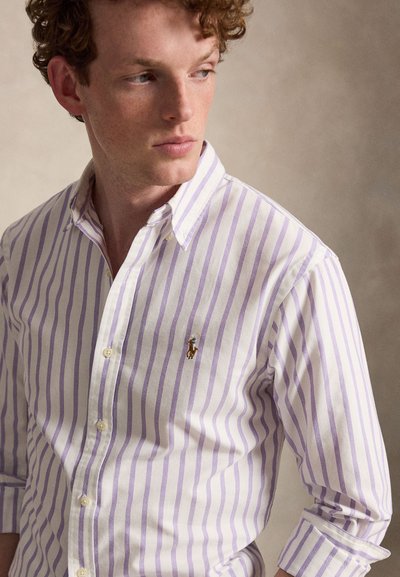 Junger Mann mit lockigem Haar, der ein weißes und hellviolettes, vertikal gestreiftes Hemd mit kleinem gesticktem Logo auf der Brust trägt.