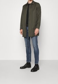 Olivgrön lång kappa med rak skärning, bärs över en svart skjorta, kombineras med blå jeans och svarta slip-on skor.