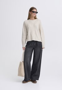 Béžový pletený svetr s texturovaným diamantovým vzorem, kombinovaný s volně střiženými tmavými džínami. Doplňky zahrnují béžovou tašku a sluneční brýle.