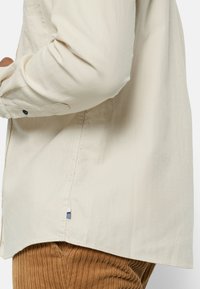 Beige button-up shirt with herringbone texture, long sleeves, and a small blue and white logo at the hem.