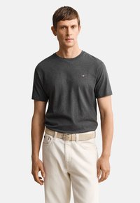 Mann trägt ein dunkelgraues T-Shirt mit Rundhalsausschnitt und kleinem Logo sowie cremefarbene Hose mit beigem Gürtel, steht vor einem schlichten Hintergrund.