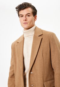 Abrigo de lana marrón con cuello de muesca, dos botones y bolsillos de solapa, superpuesto a un suéter de cuello alto color crema, de textura suave.