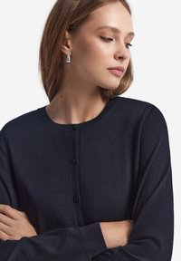 Marinblå stickad kofta med rund halslinning, långa ärmar och framknäppning. Slät textur med svarta knappar som accent.