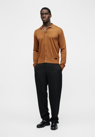 Άνδρας που στέκεται με τα χέρια στις τσέπες, φορώντας καφέ πλεκτή ζακέτα με κουμπιά, μαύρο παντελόνι και μαύρα παπούτσια επίσημου τύπου σε απλό φόντο.