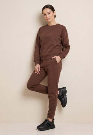 Kvinde læner sig op ad væggen iført matchende brun sweatshirt og joggingbukser med sorte sneakers, højre hånd i lommen, venstre ben bøjet.