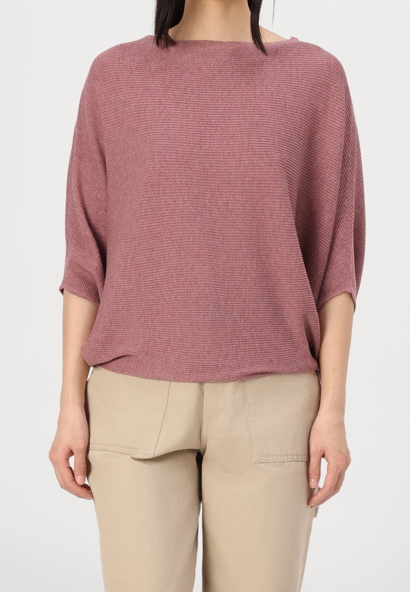Femme portant un pull ample en côtes, rose poudré à manches courtes, et un pantalon beige avec de grandes poches devant, debout devant un fond uni.