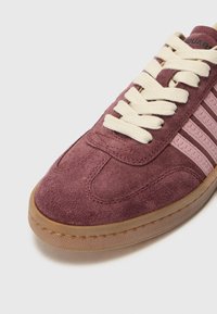 Zapatillas de ante burdeos con cordones color beige claro, que presentan franjas de acento rosas y una suela de goma texturizada, destacando un diseño atlético clásico.