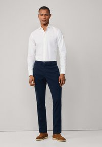 White button-up shirt with a collar, paired with navy trousers and brown suede shoes. Simple design with no visible patterns or accents.