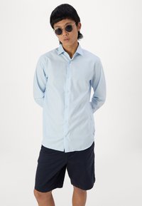 Lichtblauw geruit overhemd met lange mouwen en een kraag, gecombineerd met donkerblauwe shorts. Model draagt ronde zonnebril.
