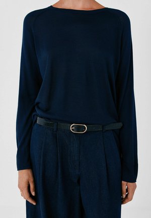 Persona che indossa una maglia a maniche lunghe blu scuro infilata in pantaloni blu scuro con una cintura nera con fibbia ovale argentata.
