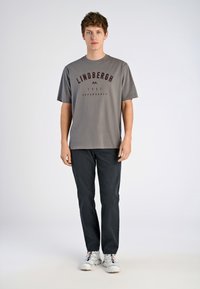 T-shirt en coton gris avec le texte maron "LINDHBERGH" et une impression graphique, associé à un pantalon sombre et des chaussures de sport de couleur claire.