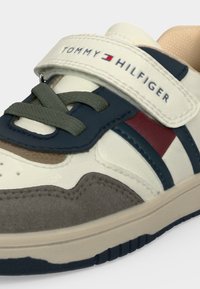 Крупен план на маратонка Tommy Hilfiger с бели, тъмносини, червени и сиви панели, тъмни връзки и каишка с велкро с марка.