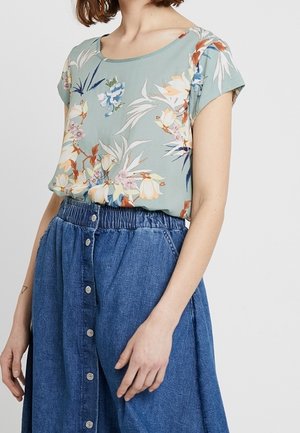 Blomstret kortærmet bluse i lys grøn, med rund halsudskæring. Sammen med en blå denimnederdel, knaplukning fortil og elastisk talje.