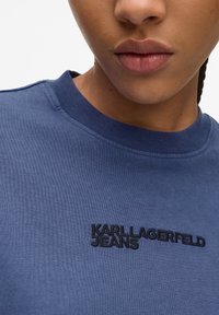 Zila kokvilnas T-kreklu ar apaļu kaklu, uz kura priekšpuses ir melni izšūti vārdi "KARL LAGERFELD JEANS", gluda tekstūra un standarta piegriezums.