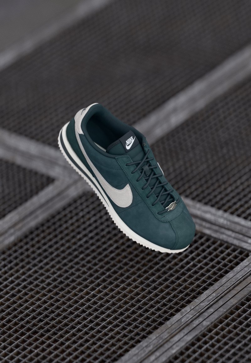 Nike αθλητικά παπούτσια σε σκούρο πράσινο σουέτ με γκρι λεπτομέρειες, διαθέτοντας στρογγυλή μύτη, επίπεδα κορδόνια και υφασμάτινη ελαστική σόλα. Λογότυπο στην γλώσσα.