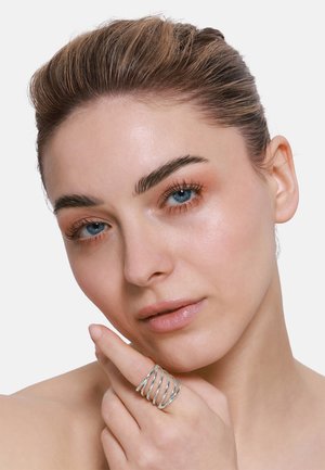 Anello a spirale argento su un dito, con molteplici bande intrecciate. La pelle del modello è liscia con trucco naturale e uno sfondo neutro.