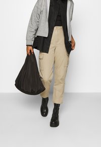 Personne marchant portant un pantalon cargo beige, des bottes noires, un sweat à capuche gris zippé par-dessus un t-shirt noir, tenant un grand sac noir dans une main.