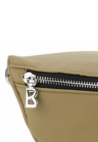 Funda de nailon beige con una cremallera negra y herrajes plateados. El tirador de la cremallera presenta una letra "B" metálica. Textura suave, forma triangular.