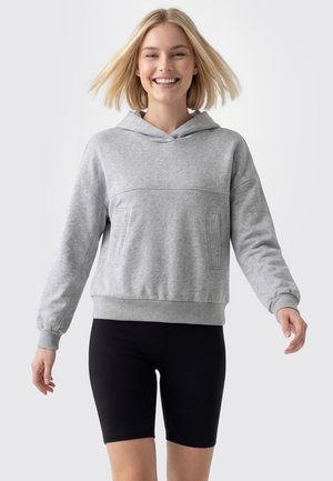 Uśmiechnięta młoda kobieta o blond włosach nosi szarą bluzę z kapturem i czarne spodenki kolarskie na jednolitym jasnym tle.