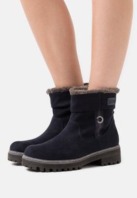 Bottes chevilles en daim bleu marine avec un revers en fausse fourrure grise texturée, semelle en caoutchouc et détail décoratif en argent sur le côté.