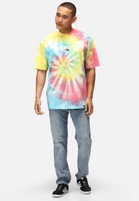 T-shirt med batikmönster i pastellfärger (gul, rosa, blå) med ett cirkulärt mönster, med texten "PATRIOTS". Bärs med ljusblå jeans och svarta sneakers.