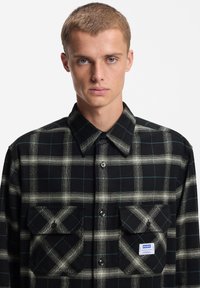 Πουκάμισο από φανέλα με ασπρόμαυρο καρό σχέδιο, μεγάλο τσέπες στο στήθος, κουμπωτό και με μπλε ετικέτα επωνυμίας. Διαθέτει κλασικό γιακά και υφή υφάσματος.