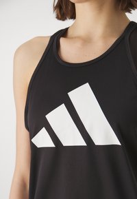 Femme portant un haut sans manches noir avec un grand logo Adidas blanc à l'avant, montrant les épaules et le haut de la poitrine.