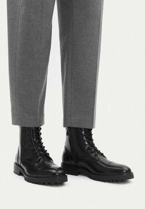 Schwarze Leder-Stiefeletten mit Schnürung, getragen zu grauen, maßgeschneiderten Hosen, dargestellt vor einem schlichten hellen Hintergrund.