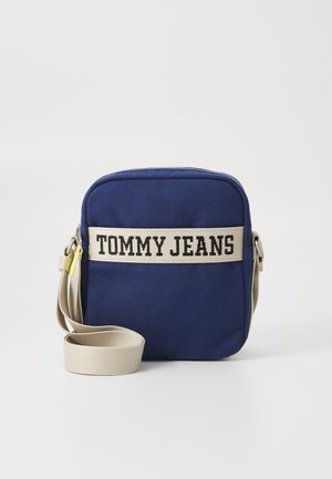 Lille firkantet navyblå crossbody-taske med beige justerbar rem og ""TOMMY JEANS"" trykt foran med sorte bogstaver på en beige rem.