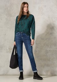 Dlouhý rukáv, tmavě zelený top s abstraktním vzorem, v kombinaci s modrými džínami, černými kotníkovými botami a černou tote taškou. Volný outfit na neutrálním pozadí.