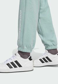 Biele tenisky s čiernymi pruhmi a logom, spárované so svetlo mentolovými teplákmi s elastickými manžetami a bočným logom značky. Textúrovaný, športový vzhľad.