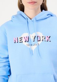 Šviesiai mėlynas džemperis, pagamintas iš minkštos medžiagos, su grafika, kurioje užrašyta "NEW YORK" ryškiomis rožinėmis ir juodomis raidėmis balto širdies dizaino fone.
