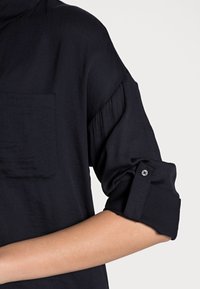 Camicia blu scuro con taschino, maniche arrotolate e un bottone decorativo. Il tessuto appare liscio con una leggera lucentezza e una consistenza morbida.