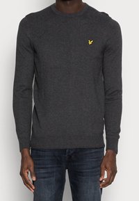 Tummanharmaa neulepaita, jossa on pyöreä pääntie, resorit hihansuissa ja helmassa, ja pieni keltainen logo rinnassa, käytetty sinisten farkkujen päällä.