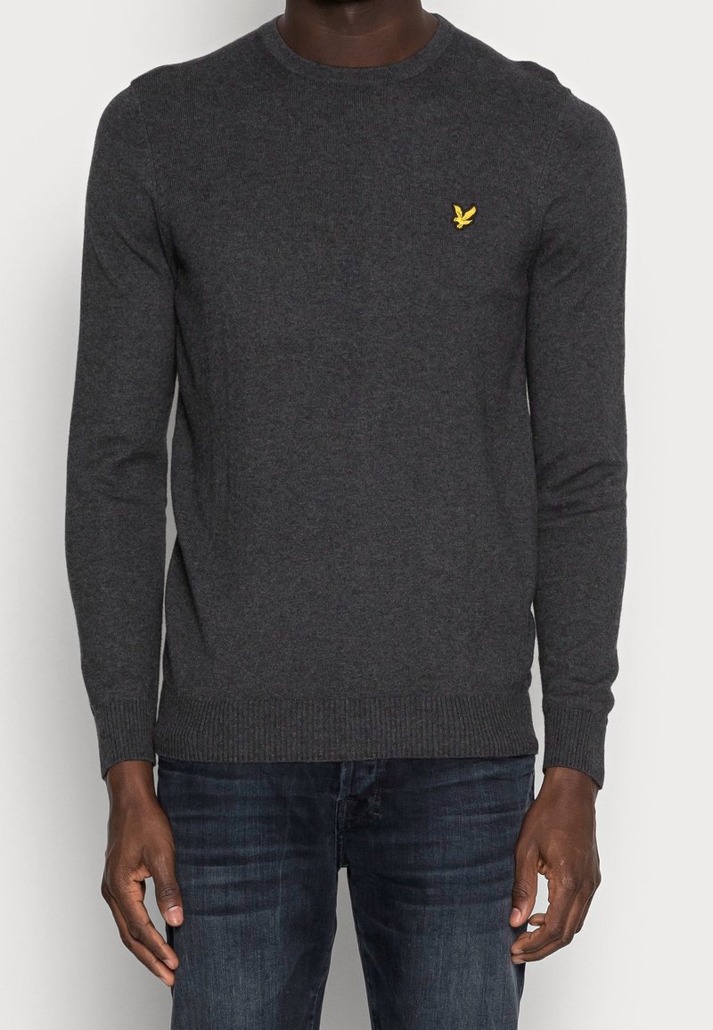 Tummanharmaa neulepaita, jossa on pyöreä pääntie, resorit hihansuissa ja helmassa, ja pieni keltainen logo rinnassa, käytetty sinisten farkkujen päällä.