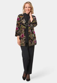 Schwarze, mit floralen Stickereien verzierte Jacke mit lockerer Passform, Dreiviertelärmeln und Knöpfen. Kombiniert mit schwarzen Hosen und hohen Schuhen.