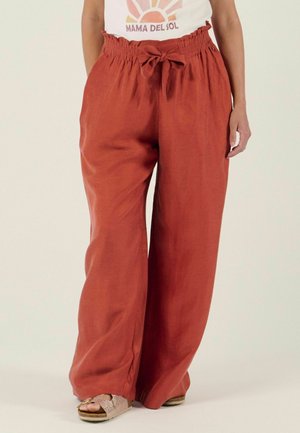 Femme portant un pantalon large ample couleur rouille orange avec une taille élastique et un nœud, associé à des sandales métalliques plates et un haut graphique.