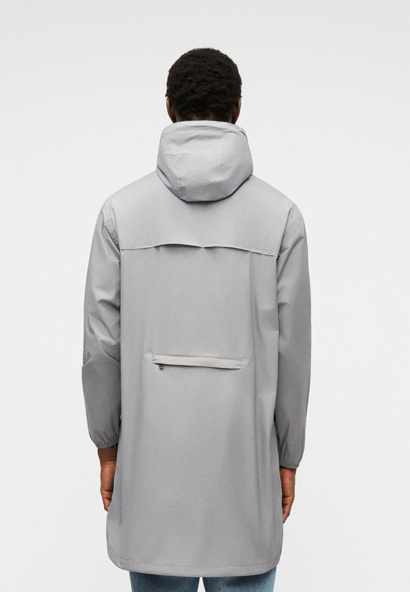 Homme portant un imperméable long gris clair avec capuche, poignets elastiques et poche arrière zippée, vu de dos.