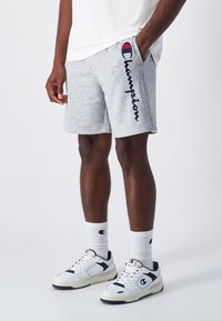 Graue Baumwollshorts mit in Marineblau gesticktem "Champion", Seitentaschen, kombiniert mit weißen Socken, die ein Logo zeigen, und weißen/schwarzen Sportschuhen.