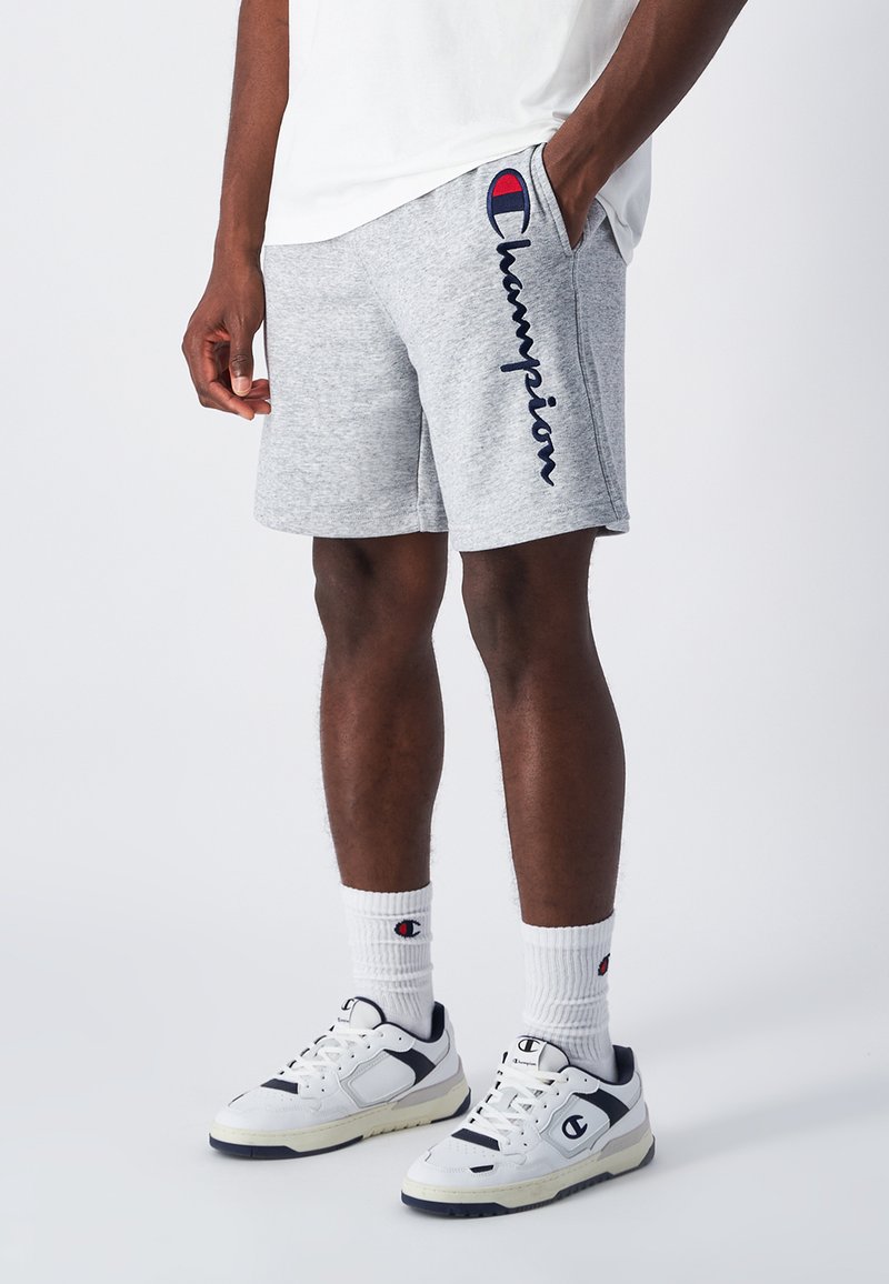 Graue Baumwollshorts mit in Marineblau gesticktem "Champion", Seitentaschen, kombiniert mit weißen Socken, die ein Logo zeigen, und weißen/schwarzen Sportschuhen.