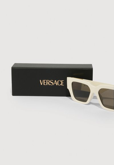 Kremovo sfarbené obdĺžnikové slnečné okuliare Versace s tmavými šošovkami, ležiace vedľa čiernej krabičky Versace so zlatým písaním.